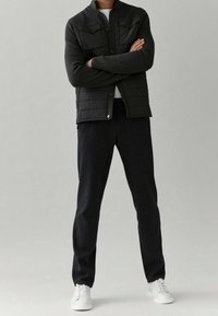 Black quilted jacket with snap buttons and two chest pockets, paired with dark trousers and white sneakers. Smooth texture and fitted design.
