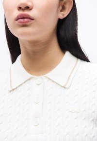 Κοντινό πλάνο ενός ατόμου που φοράει λευκό πόλο πουλόβερ με πλεκτό σχέδιο καλωδίου, κουμπιά και μικρό κεντημένο λογότυπο στο στήθος.