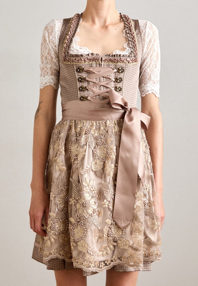 Femme portant une robe dirndl beige avec des manches en dentelle, une broderie florale, un nœud en ruban de satin et des fermoirs métalliques décoratifs sur le corsage.