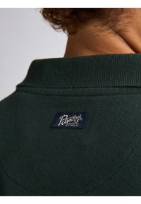 Donkergroen overhemd met kraag en een gestructureerde stof, met een marineblauw label met witte letters op de achterhals.