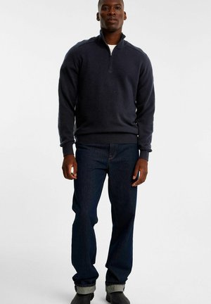 Mørk navy sweater med en lynlåset krave, parret med mørkeblå jeans. Sweateren har ribbede ærmer og kant, med en glat tekstur.