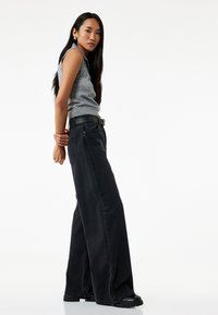 Jeans negros de tiro alto y pierna ancha con dobladillos deshilachados y detalles en los bolsillos delanteros, combinados con una camiseta sin mangas ajustada gris.
