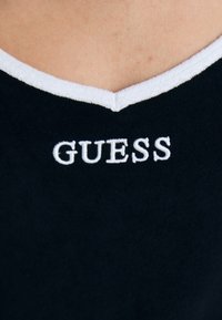 Camisa negra con escote en V blanco y logo bordado "GUESS" en blanco en la parte delantera. El tejido parece suave y texturizado.