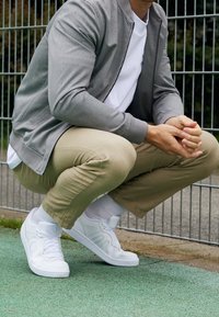 Grå bomberjacka för män över en vit t-shirt, kombinerad med beige byxor och vita sneakers. Scenen inkluderar en grön utomhusyta och stängsel.