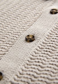 Knit cardigan in beige with textured pattern. Features dark brown buttons along the front. Close-up view highlights the material's details.