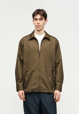 Młody mężczyzna w oliwkowo-zielonej bluzie zapinanej na zamek, na białej koszuli i ciemnych spodniach, stoi na jednolitym jasnym tle.