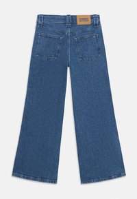 Jean large en denim bleu avec une finition texturée, doté de deux poches arrière et d'une patch en cuir marron portant l'inscription "TOMMY HILFIGER" sur la ceinture.