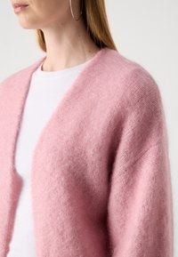Cardigan rosa pallido realizzato in materiale morbido e peloso, con una vestibilità comoda e design aperto, abbinato a una t-shirt bianca sottostante.