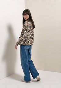 Trui met luipaardprint in beige en bruin, gecombineerd met blauwe wijde jeans. Het model staat tegen een lichte achtergrond en kijkt achterom.
