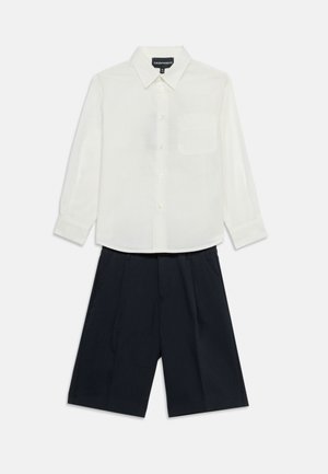 Cămașă albă cu mâneci lungi, cu nasturi și buzunar la piept, asortată cu pantaloni scurți croiți, de culoare bleumarin închis, pe un fundal alb.