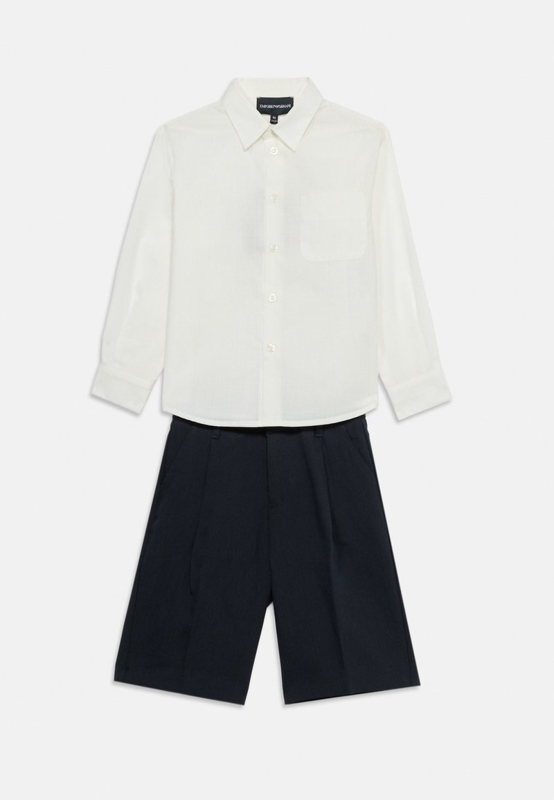 Άσπρο πουκάμισο με μακριά μανίκια και κουμπιά, με τσέπη στο στήθος, συνδυασμένο με σκούρο μπλε σορτς ραμμένο στο μέτρο, σε λευκό φόντο.
