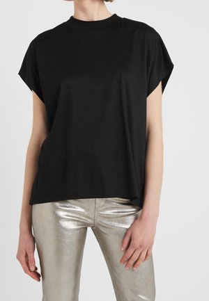 Personne portant un haut noir ample à manches courtes et un pantalon argenté métallisé ajusté, debout devant un fond clair uni.