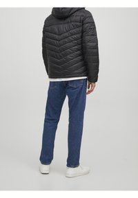 Hombre con chaqueta acolchada negra, jeans azules y zapatillas blancas, de pie mirando hacia fuera contra un fondo claro y liso.