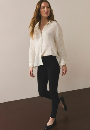 Naine seisab neutraalse taustaga, kandes lahtist valget nööpidega pluusi, musti vormitud pükse ja musti korterjalatseid, vaadates küljele.