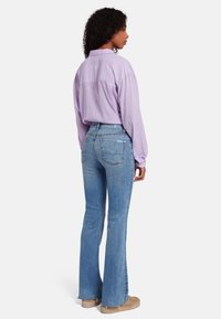 Chemise lilas à col, à manches longues, associée à un jean flare bleu délavé. La tenue présente des coutures discrètes et une coupe décontractée.