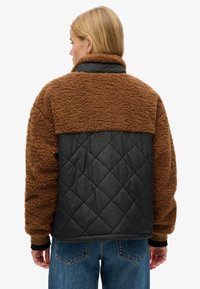 Giacca marrone pelosa con retro nero trapuntato, caratterizzata da un colletto alto e maniche lunghe. La texture è morbida e vellutata.