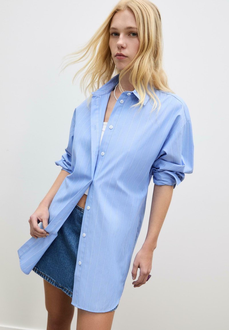 Ung kvinna med långt blont hår bär en lös, ljusblå randig skjorta över en vit topp och en denim mini-kjol mot en neutral bakgrund.