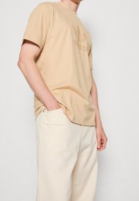 Camiseta de algodón beige con un logo de Adidas texturizado en marrón, combinada con pantalones de algodón en beige claro. Corte relajado, mangas cortas.