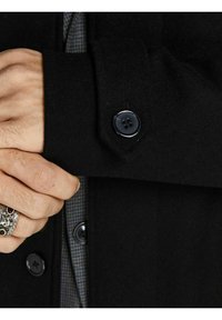 Svart ylleblandad kappa med knappärmar, som visar en texturerad yta och en skjorta med dolda rutor under.