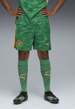 Completo calcistico verde con un motivo a gradiente testurizzato. Pantaloni corti con dettagli arancioni e logo della squadra. Calze lunghe fino al ginocchio in verde, con la scritta "CITY" e il logo.