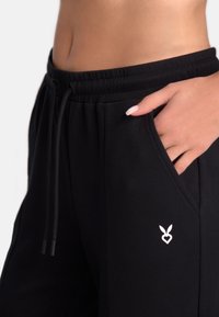 Pantaloni de jogging negri, confecționați dintr-un material moale, au o talie elastică cu șnur și buzunare laterale, evidențiate de un mic logo alb.