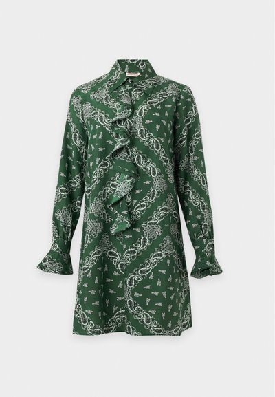 Πράσινο μπλουζάκι με μακριά μανίκια με άσπρο μοτίβο παϊλέι, βολάν στο μπροστινό μέρος και μυτερό γιακά σε μονόχρωμο φόντο.