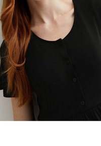 Detailní záběr osoby v černém topu s knoflíkovým výstřihem, jejíž dlouhé, vlnité červené vlasy rámují její obličej a krk.