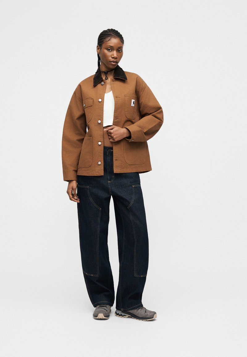 Donna con i capelli intrecciati che indossa una giacca marrone con bottoni, un top bianco corto, jeans larghi scuri e sneakers grigie, in piedi davanti a uno sfondo semplice.