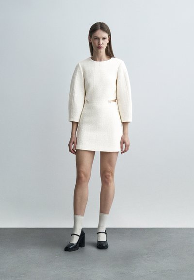 Γυναίκα που φορεί ένα λευκό μίνι φόρεμα με υφή, με μακριά φουσκωτά μανίκια και κοψίματα στα πλάγια, συνδυασμένο με λευκές κάλτσες και μαύρα τακούνια Mary Jane.