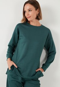 Türkiz hosszú ujjú pulóver, puha anyagból készült, kerek nyakkal, ejtett vállakkal és oldalsó hasítékokkal az alján.