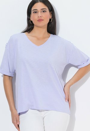 Kvinde iført lyse lilla V-hals kortærmet top med knapdetaljer på skuldrene, hvide bukser og gyldne hoop-øreringe, stående med hånden på hoften.
