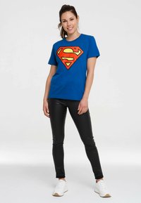 Blaues Baumwoll-T-Shirt mit einem großen roten und gelben Superman-Logo, kombiniert mit schwarzen Leggings und weißen Turnschuhen. Einfacher Rundhalsausschnitt.
