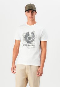 Jaunas vyras, vilkintis baltu marškinėlių su pilku piešiniu ir tekstu „BELSTAFF“, kreminėmis kelnėmis ir languota kepure, stovi prieš baltą foną.