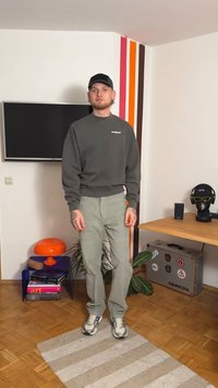 Sweatshirt cinzento, calças verdes e ténis. Em pé sobre um tapete às riscas numa sala com detalhes de parede coloridos e mobiliário.