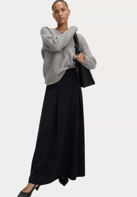 Grau gerippter Pullover mit V-Ausschnitt, kombiniert mit einem schwarzen, plissierten Maxikleid und schwarzen, spitzen Schuhen, während eine schwarze Handtasche gehalten wird.
