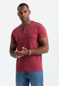 Camisa henley roja de manga corta con tres botones negros en el cuello, combinada con jeans azules. Textura suave, corte casual.