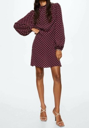 Femme portant une robe bordeaux à manches longues avec des pois blancs et des talons hauts nude à brides, debout avec un bras plié à la taille.