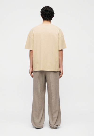 Άτομο με σγουρά σκουρόχρωμα μαλλιά, στραμμένο με την πλάτη, φοράει ένα φαρδύ μπεζ πουκάμισο με κοντά μανίκια και φαρδιά γκρι παντελόνια, στέκεται μπροστά σε απλό φόντο.