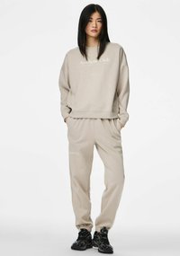 Beżowy komplet z bluzą i dresami z napisem "Aesthetic Club". Miękka, fakturowana tkanina, luźny krój, elastyczne mankiety w spodniach. Czarnе sneakersy dopełniają stylizację.