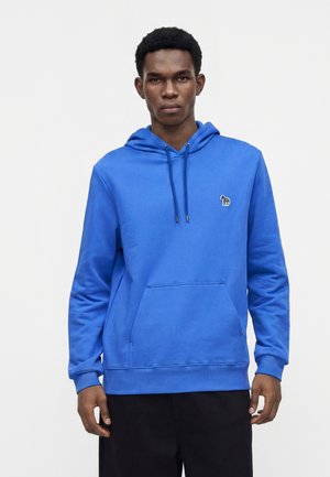 Homme portant un sweat à capuche bleu vif avec une poche avant et un petit logo zèbre sur la poitrine, debout devant un fond clair uni.
