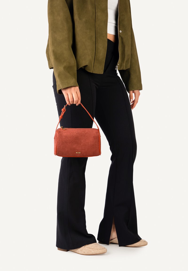 Femme portant une veste olive, pantalon noir évasé, chaussures beige, tenant un petit sac à main couleur rouille avec une poignée supérieure sur fond blanc.