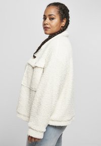 Hvit fleecejakke med en teksturert, fluffy overflate, krage og lange ermer. Passformen er løs og avslappet, kombinert med lyseblå jeans.