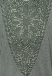 Besticktes Detail auf einem dunkelgrünen Stoff mit einem floralen Muster, das mit strukturierten Akzenten und einem Mesh-Hintergrund versehen ist.