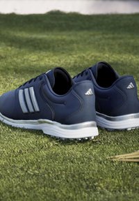 Ζευγάρι αθλητικά παπούτσια Adidas σε σκούρο μπλε χρώμα με λευκές σόλες, που ακουμπούν σε πράσινο γρασίδι έξω.
