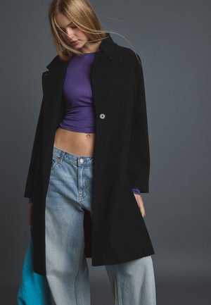 Jeune femme portant un manteau noir par-dessus un crop top violet et un jean bleu clair, regardant vers le bas avec les cheveux au vent sur un fond gris.