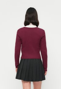 Femme aux cheveux bruns mi-longs portant un pull moulant bordeaux et une jupe plissée noire, se tenant face à un mur blanc.
