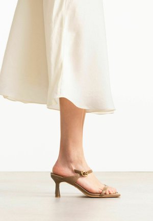 Femme portant des sandales à talons beiges avec bride et boucle à la cheville, associées à une jupe midi blanche fluide sur un fond uni.