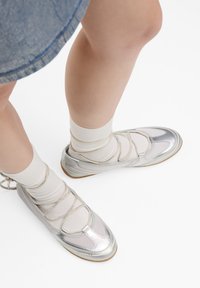 Ballerines argentées avec un mélange de mesh et de cuir verni, présentant un laçage croisé et une semelle en caoutchouc, assorties à des chaussettes blanches.