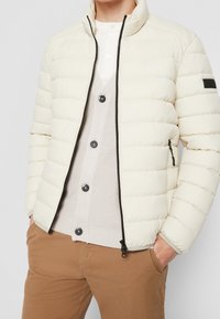 Giacca cremosa e gonfia con colletto alto, zip nera a contrasto e tasche laterali. Indossata sopra un cardigan beige chiaro e pantaloni marroni.
