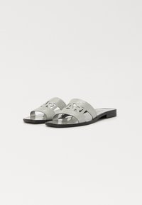 Chanclas de purpurina plateada con una suela negra plana, que presentan letras recortadas en la correa frontal y un acabado suave y brillante.